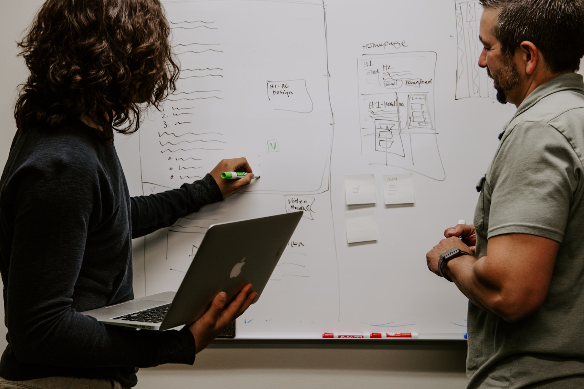 Two colleagues collaborate on a design by drawing it out on a whiteboard while holding a laptop